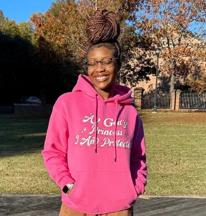 Person wearing a pink Christian hoodie with "As God's Princess I am protected", standing outdoors in a park.