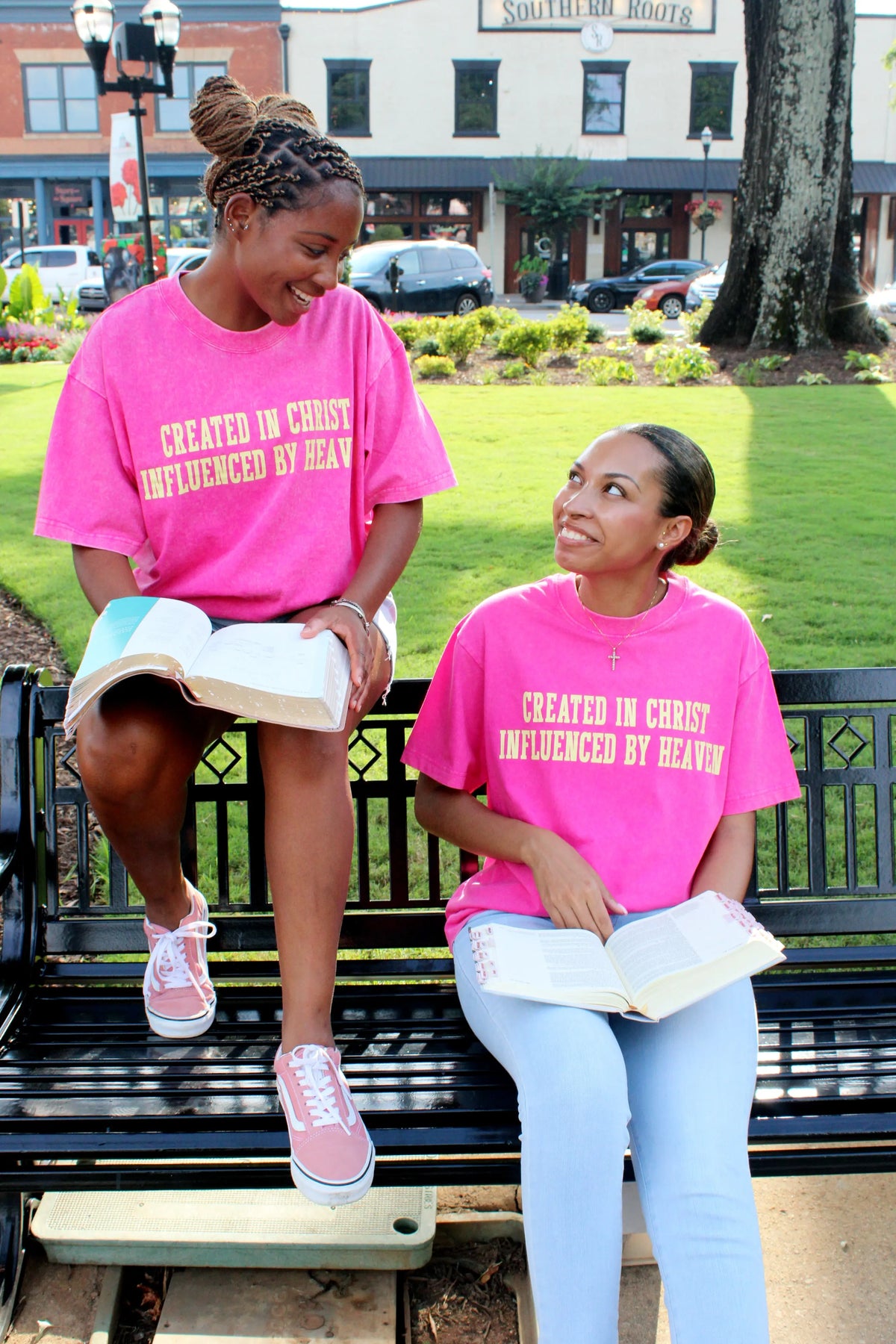 Two women sitting on a bench wearing pink Christian t-shirts with text, in an outdoor setting.